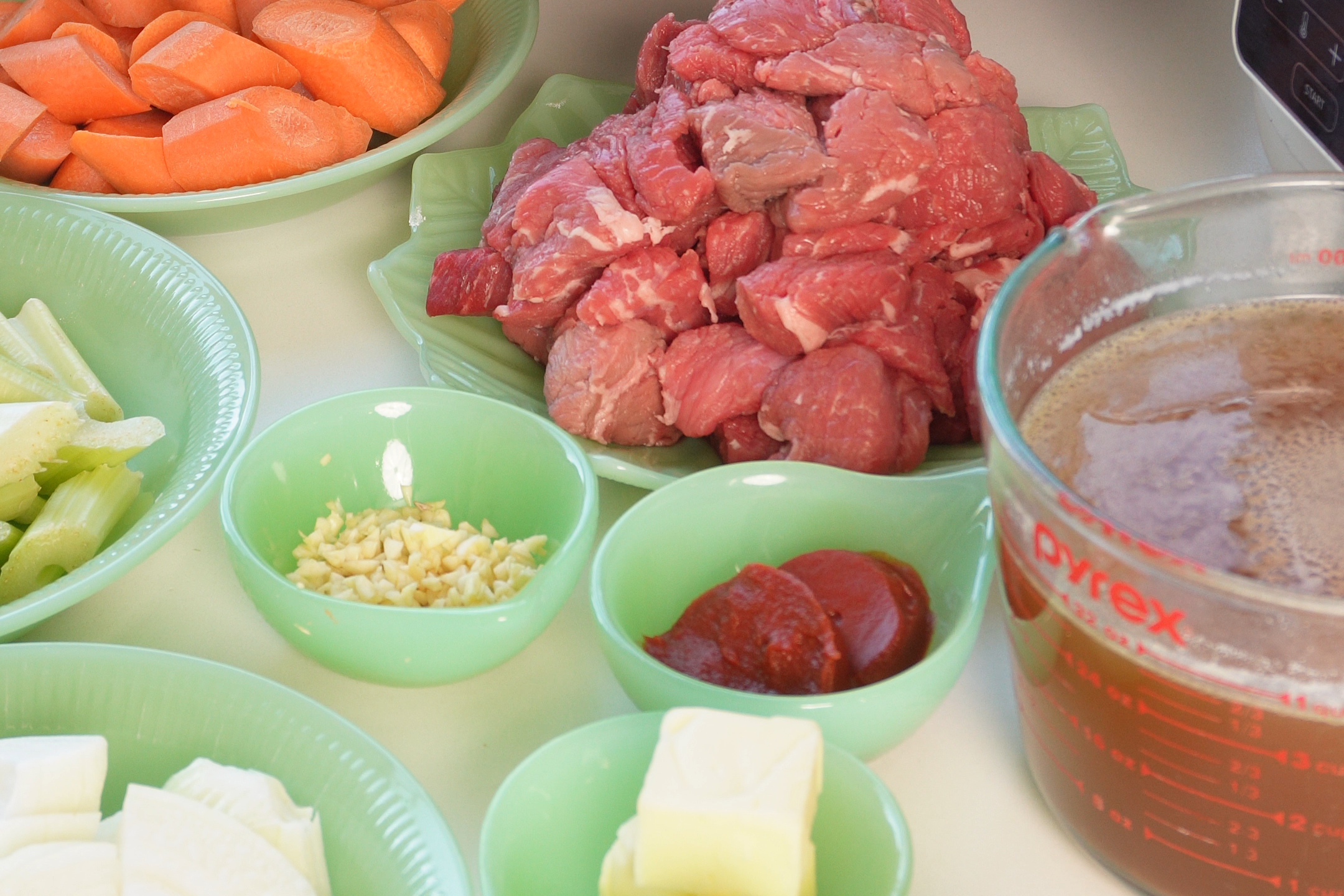 Ingredients for a meal prep including chopped potatoes, carrots, celery, onions, a pile of cubed beef, garlic, tomato paste, butter, a measuring cup of broth, and a bottle of DAngelo Aglianico del Vulture wine on a wooden stand.