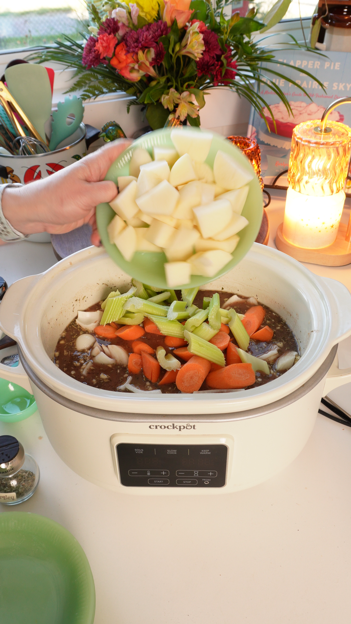A hand pours chopped potatoes from a green bowl into a Crock-Pot filled with carrots, celery, and onions in broth.