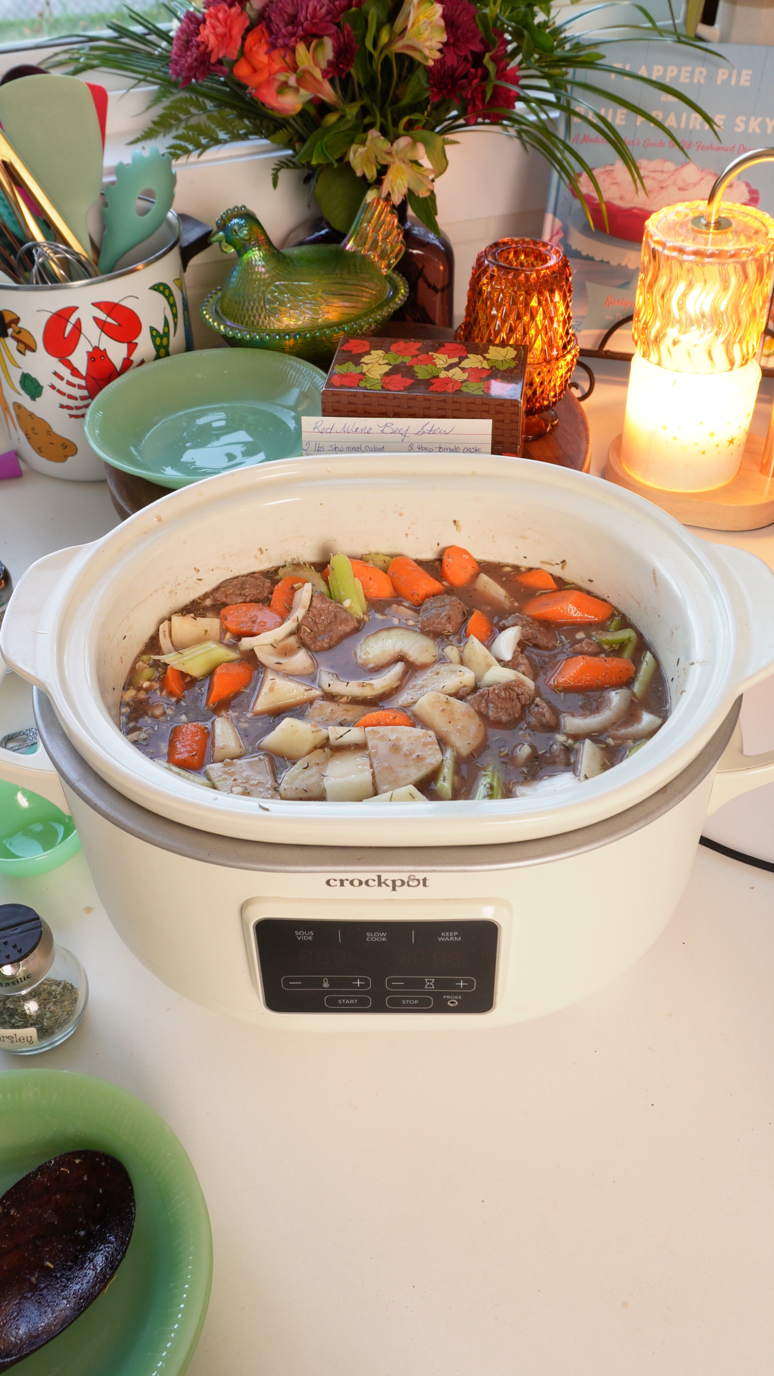 Slow cooker filled with beef stew containing carrots, potatoes, and onions, with a recipe card reading Red Wine Beef Stew visible in the background.