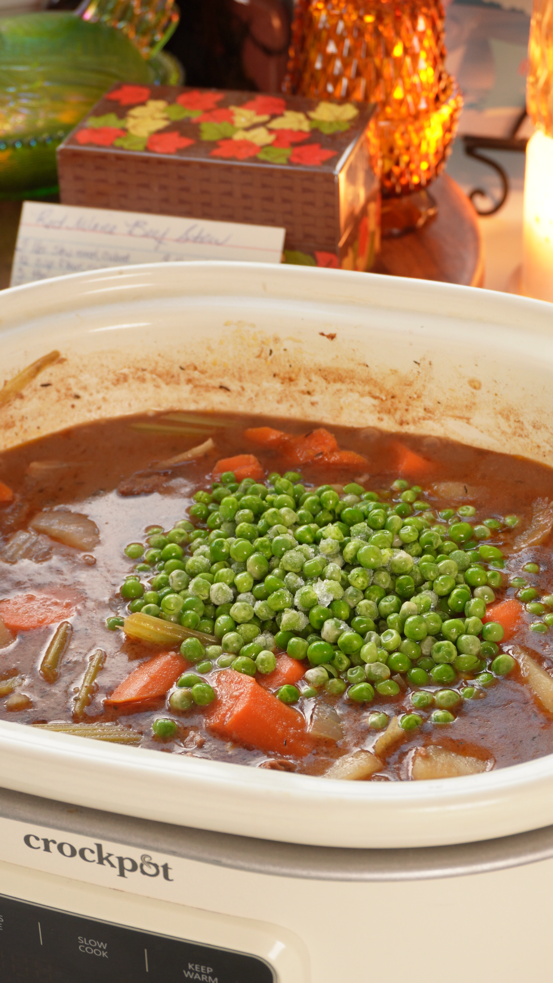 Beef stew with carrots, celery, and a pile of frozen green peas cooking in a white Crock-Pot.