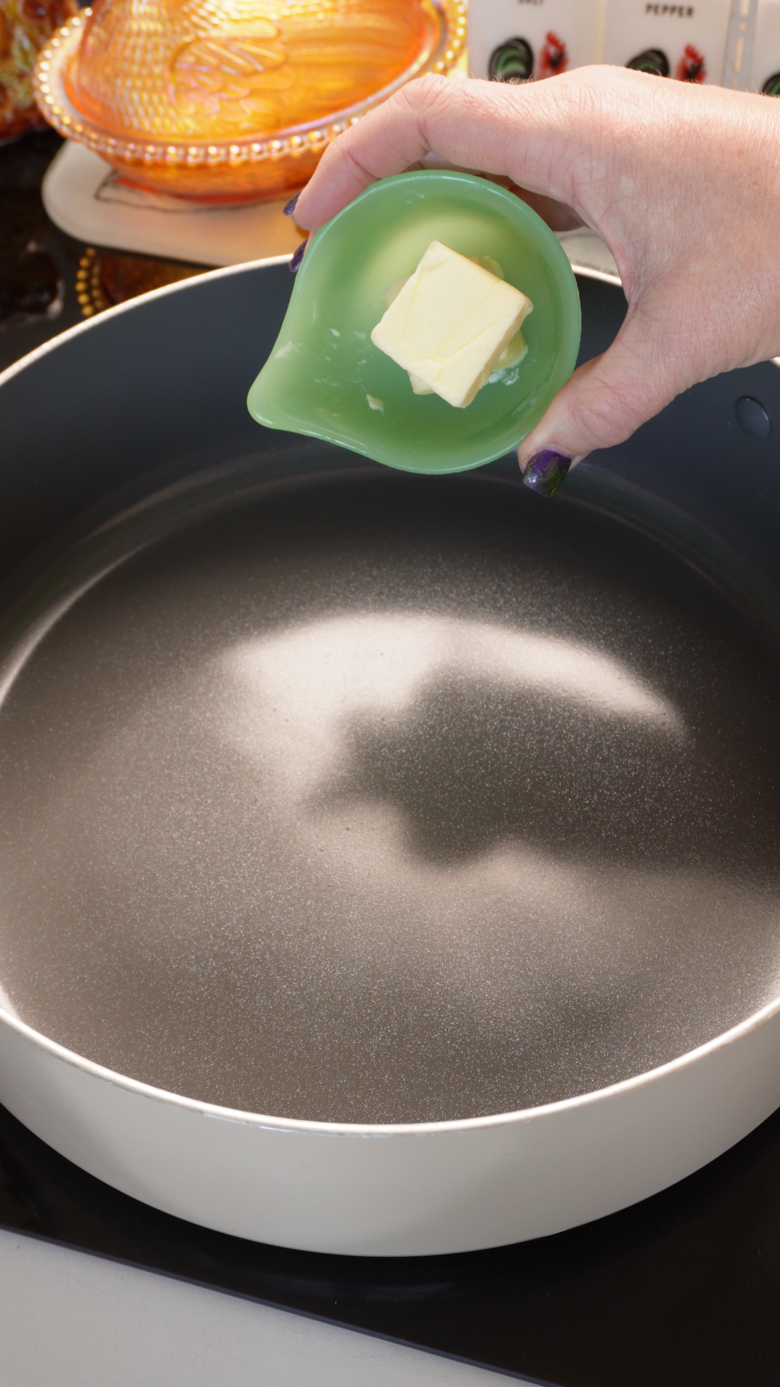 A hand holds a small green dish containing a pat of butter over an empty frying pan.