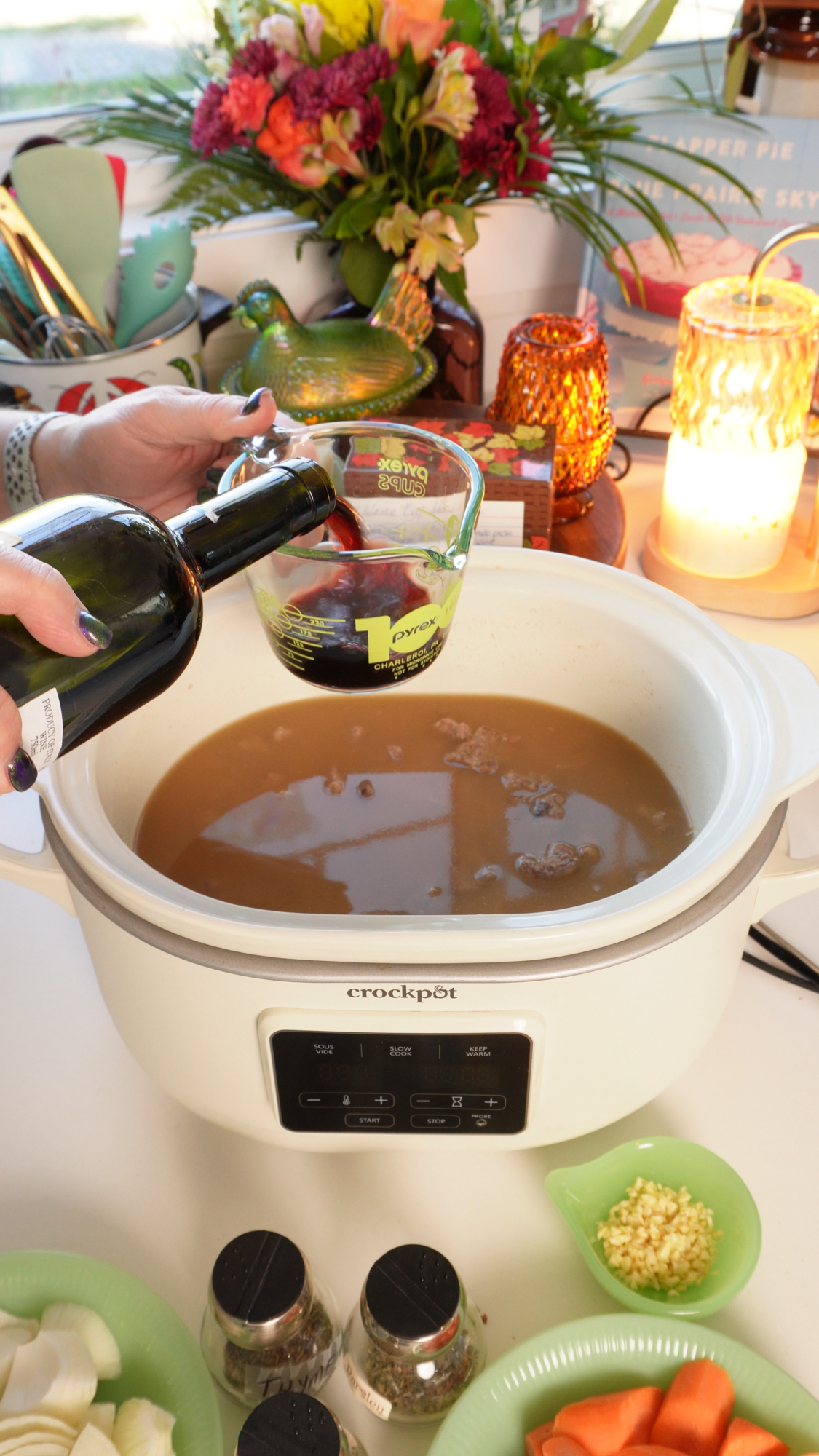 A hand pours red wine from a bottle into a Pyrex measuring cup, held over a white Crockpot containing broth and meat.