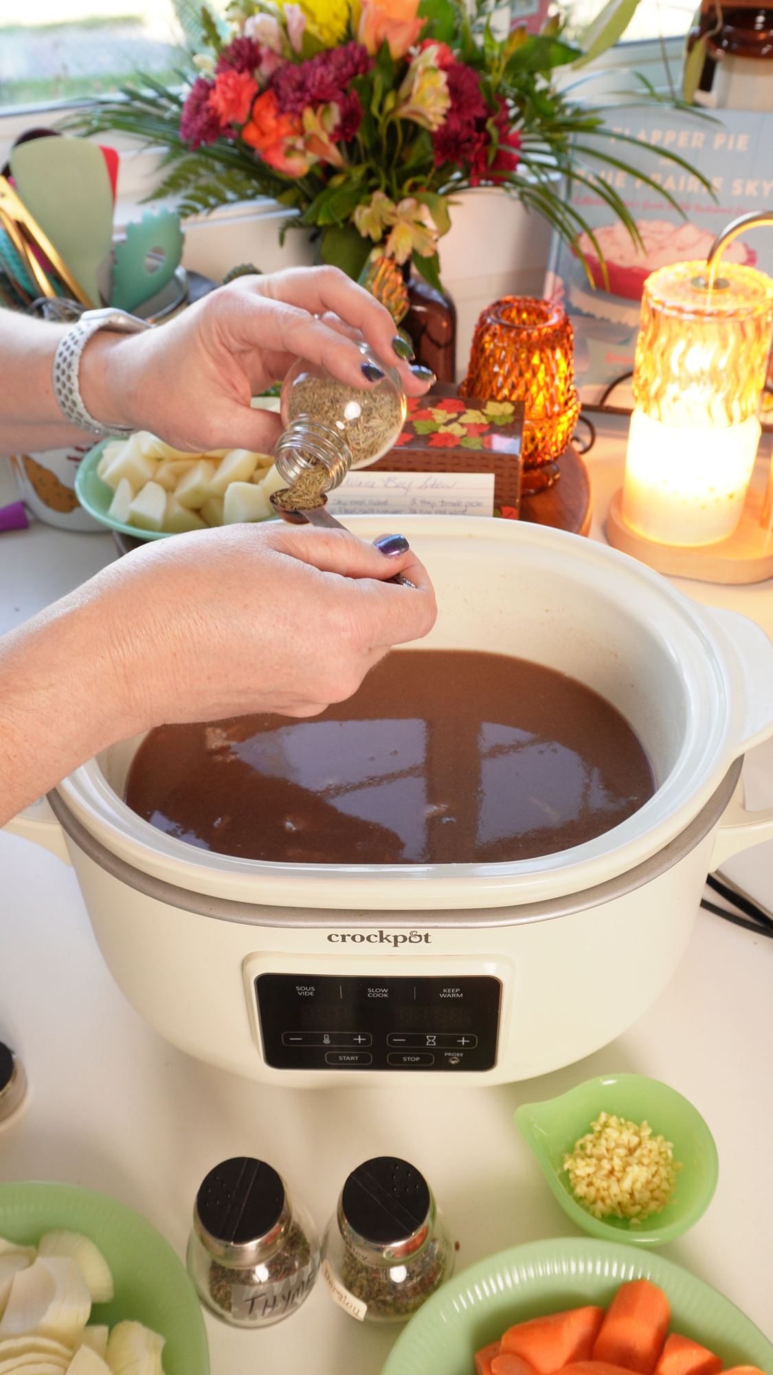 Hands pouring dried herbs from a spice jar into a measuring spoon held over a slow cooker filled with brown broth.