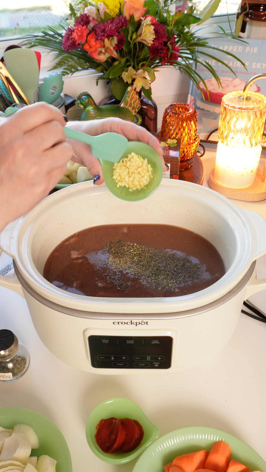 A hand holds a small green scoop of minced garlic over a slow cooker filled with brown liquid and herbs.