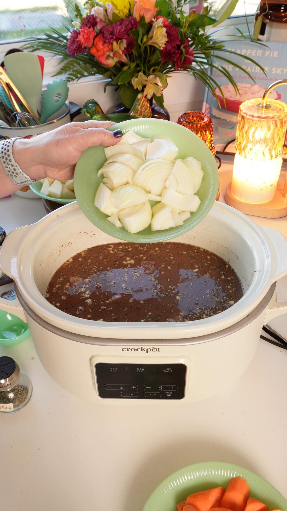 A hand holds a green plate of chopped white onions over a slow cooker filled with brown liquid.