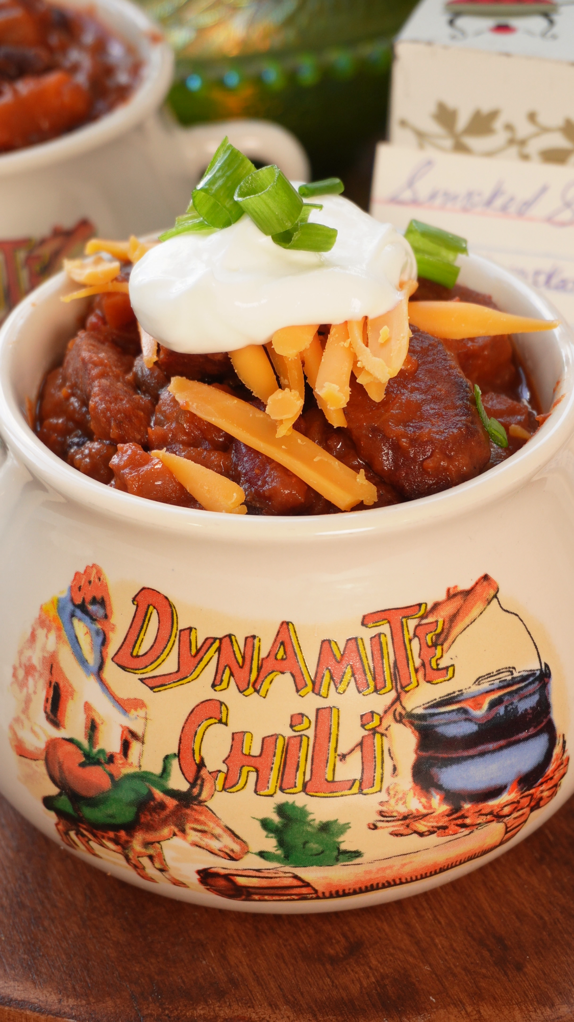 A bowl of chili topped with shredded cheddar cheese, sour cream, and sliced green onions, served in a ceramic crock labeled Dynamite Chili.