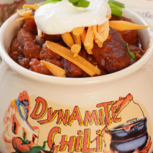 A bowl of chili topped with shredded cheddar cheese, sour cream, and sliced green onions, served in a ceramic crock labeled Dynamite Chili.