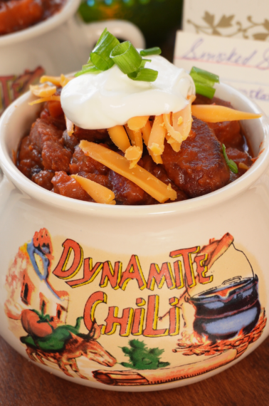 A bowl of chili topped with shredded cheddar cheese, sour cream, and sliced green onions, served in a ceramic crock labeled Dynamite Chili.