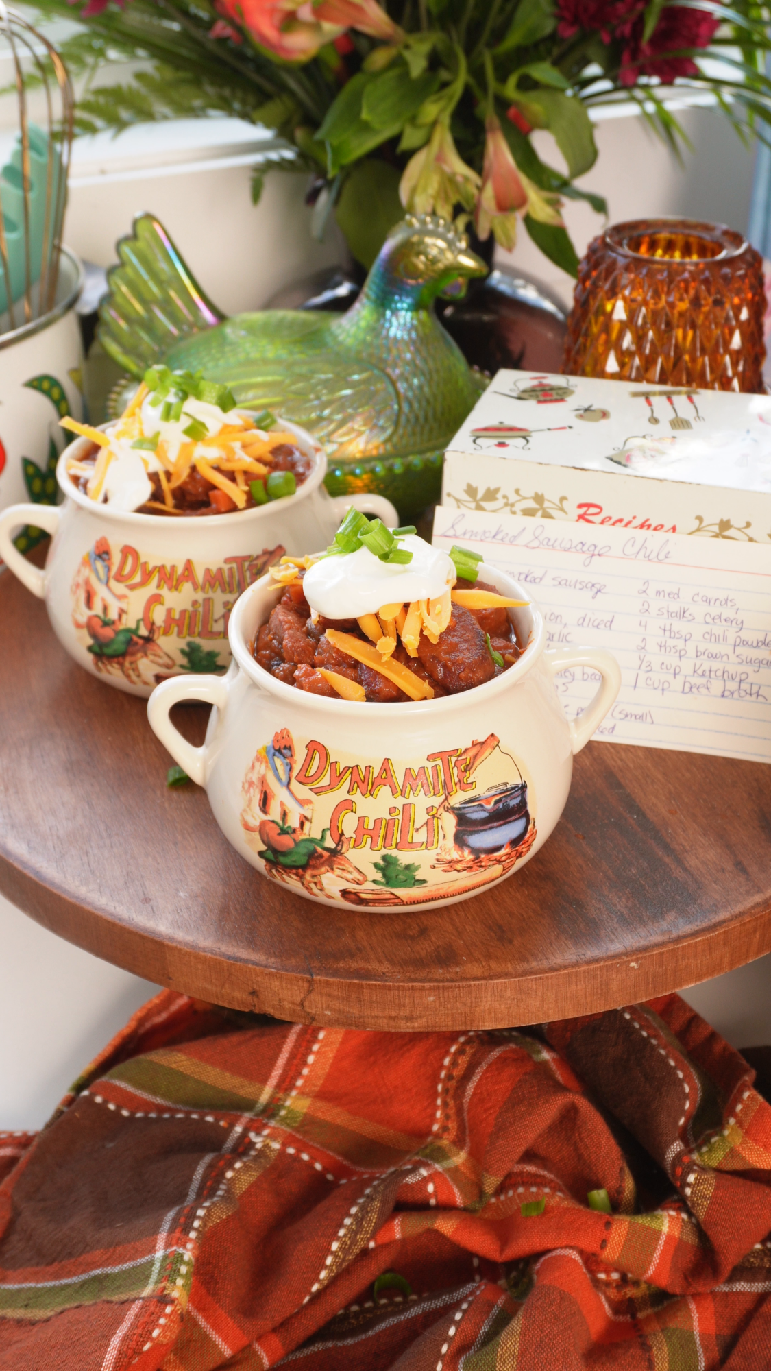 Two ceramic bowls of chili topped with shredded cheese, sour cream, and green onions, sitting on a wooden surface next to a handwritten recipe card.