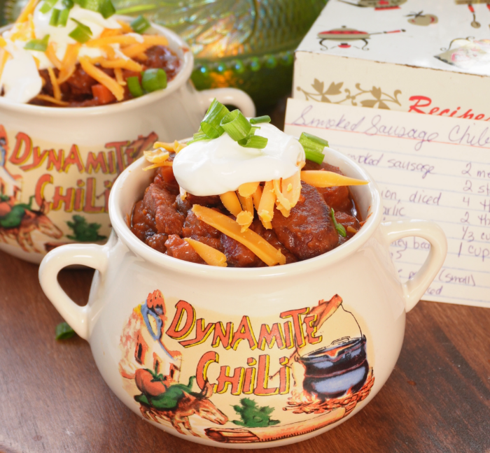 Two ceramic bowls labeled Dynamite Chili, filled with chili topped with sour cream, shredded cheese, and green onions, sitting next to a handwritten recipe card.