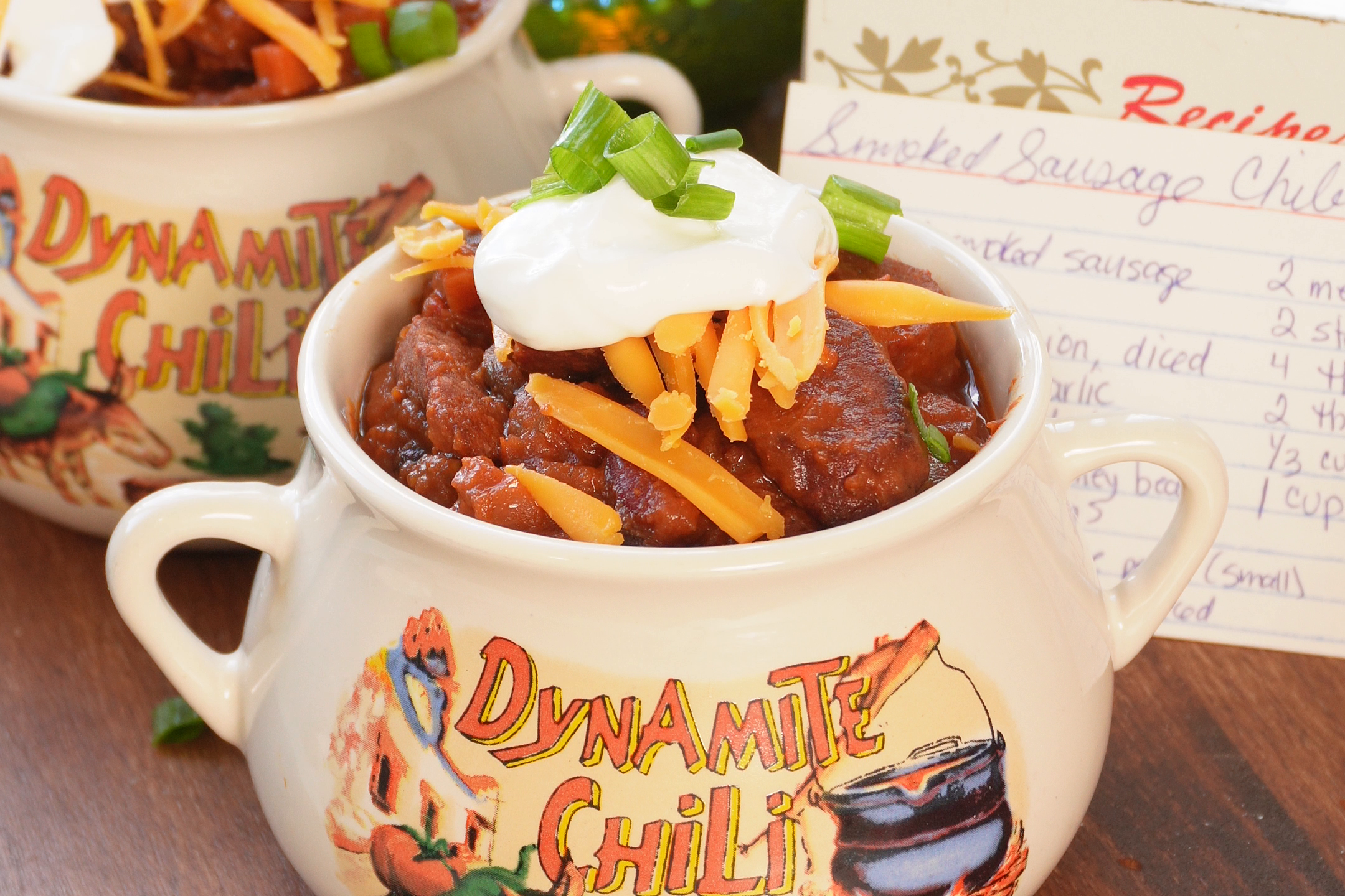 Two ceramic bowls labeled Dynamite Chili, filled with chili topped with sour cream, shredded cheese, and green onions, sitting next to a handwritten recipe card.