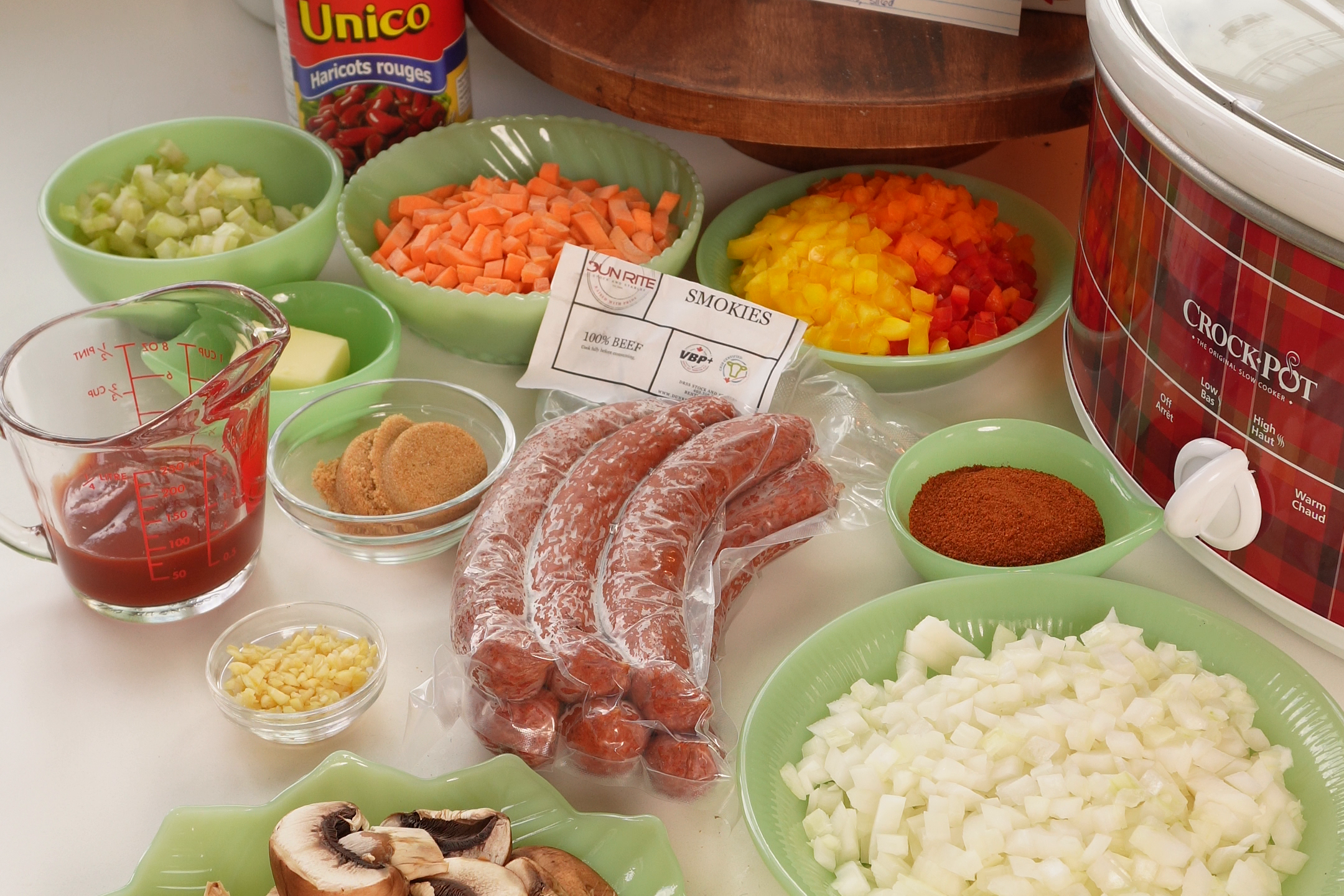 Ingredients for a slow cooker sausage chili, including chopped vegetables, mushrooms, sausages, canned beans, tomatoes, and a recipe card spread across a kitchen counter.