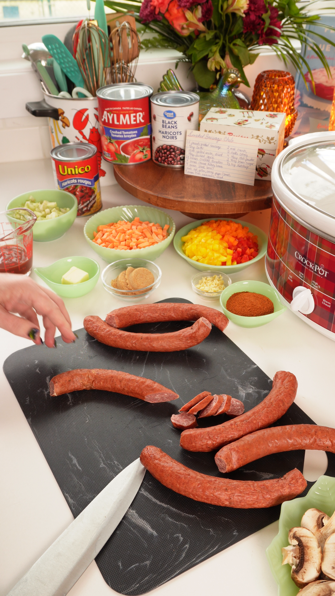 A person slicing smoked sausage on a cutting board surrounded by bowls of chopped vegetables, canned goods, and a slow cooker.