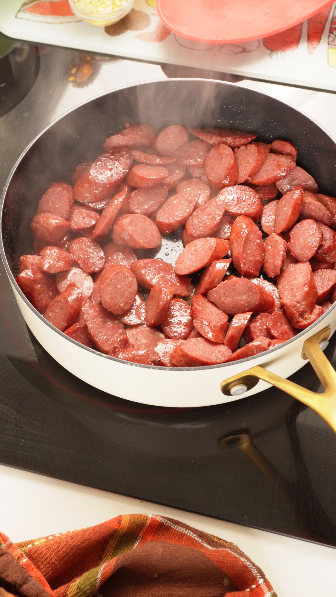 Sliced sausage cooking in a steaming white skillet on a black stovetop.