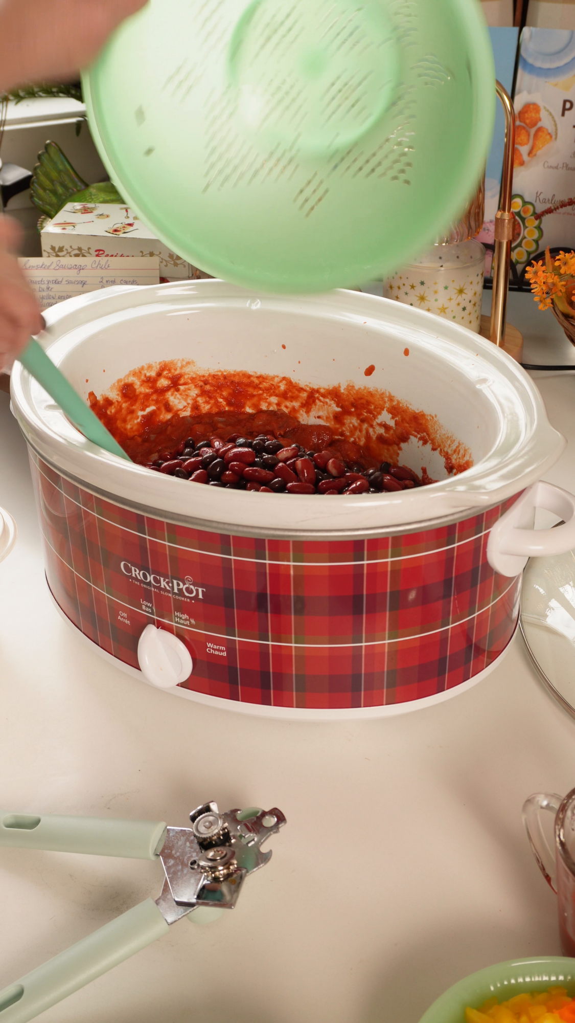 A person uses a green spatula to stir kidney beans and tomato sauce inside a plaid-patterned Crock-Pot slow cooker.