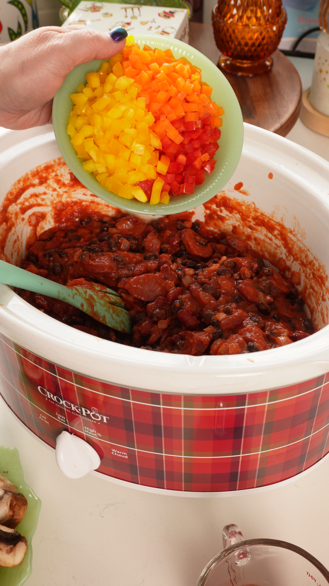 A hand pours diced yellow and orange bell peppers into a slow cooker filled with a red bean and sausage stew.