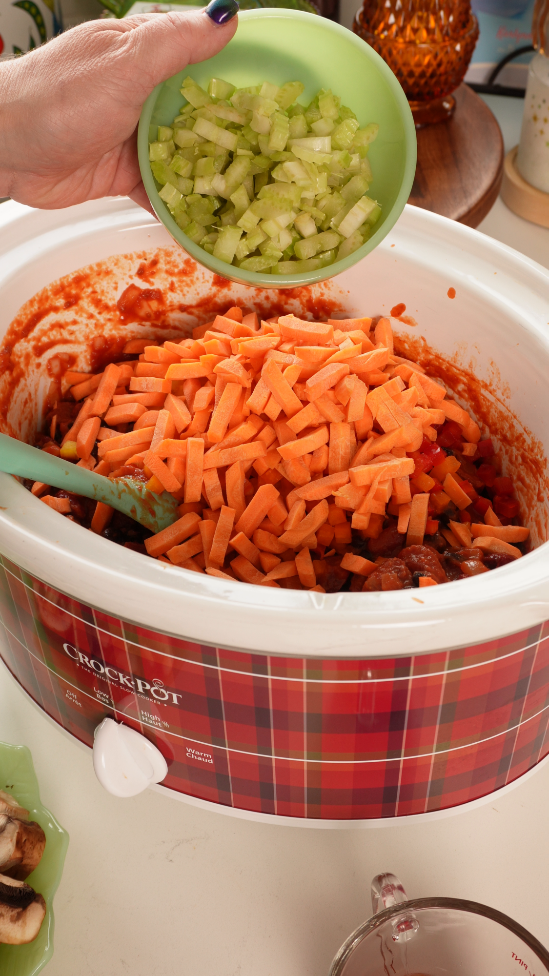 A hand pours a bowl of diced celery into a slow cooker filled with carrots and a tomato-based mixture.