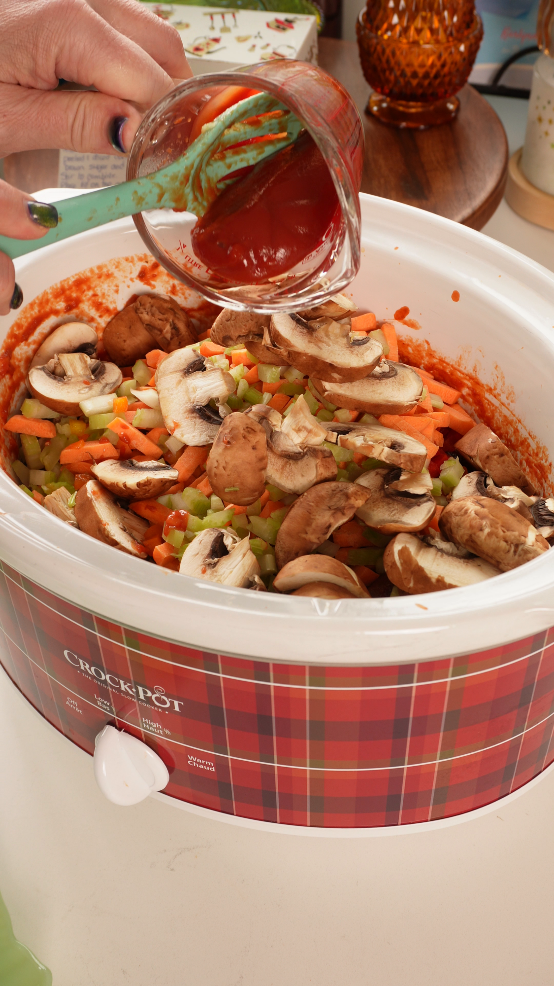 A hand pours tomato sauce from a measuring cup into a plaid Crock-Pot filled with sliced mushrooms, chopped carrots, and celery.