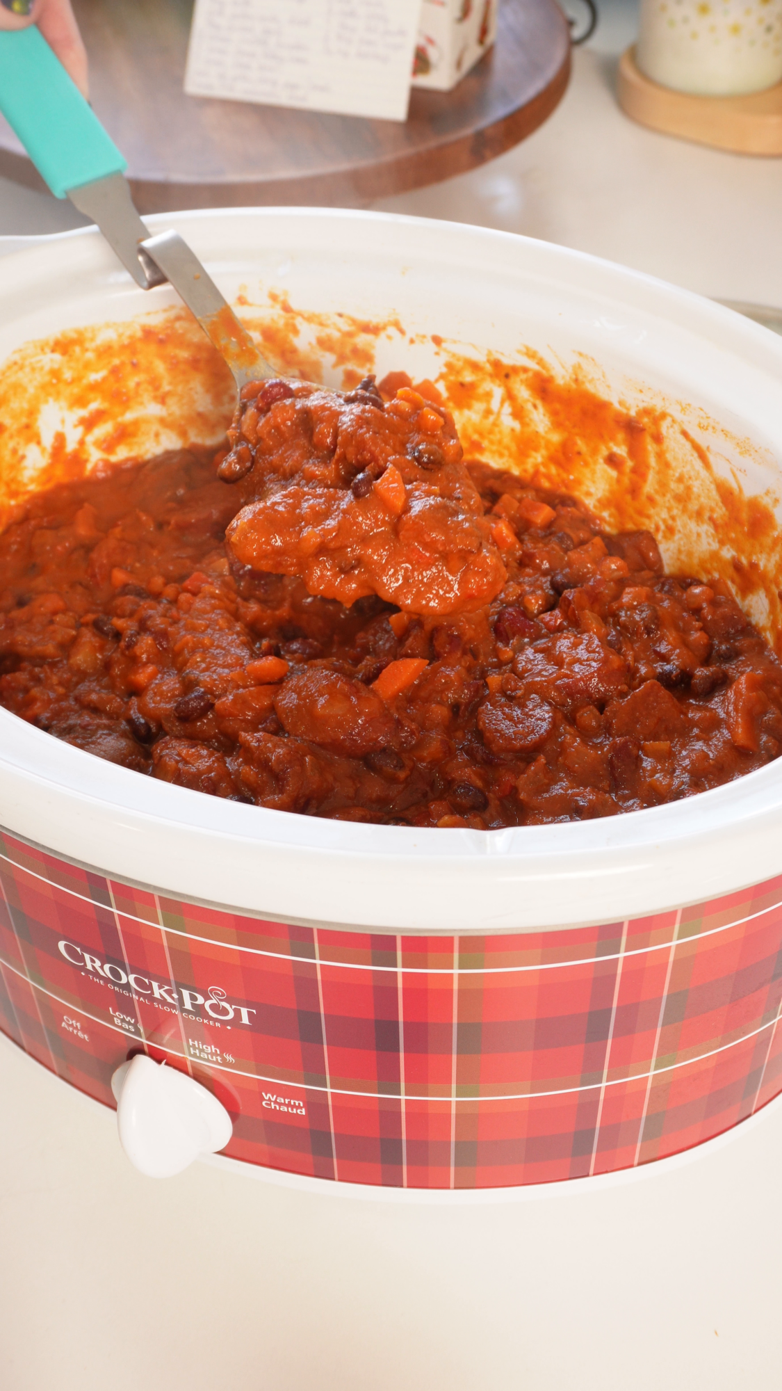 A plaid-patterned Crock-Pot filled with chunky vegetable and bean chili, being scooped with a ladle.