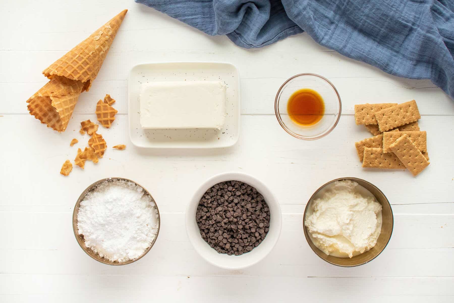 Ingredients for a dessert laid out on a white surface, including a block of cream cheese, waffle cones, graham crackers, powdered sugar, chocolate chips, a bowl of cream, and a small dish of vanilla extract.