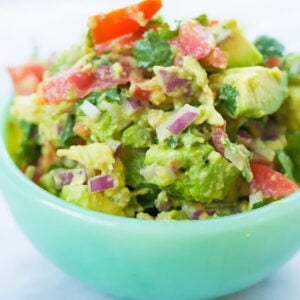 A light green bowl filled with chunky guacamole containing diced tomatoes, red onions, and cilantro.